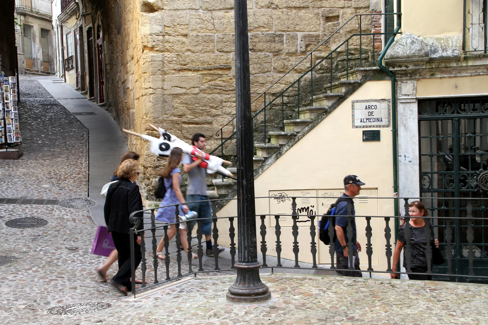 Besucher auf den Stufen neben dem Almedina-Bogen, mit einer Straßenlaterne, einem Eisengeländer und dem Namensschild des Bogens an der Wand
