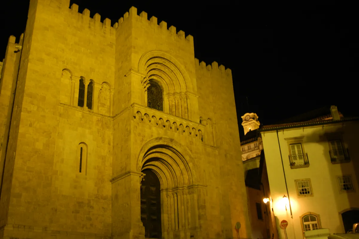 La fachada románica almenada de la Catedral Vieja de Coímbra, dorada por la luz contra el cielo nocturno, con casas iluminadas al borde de la plaza