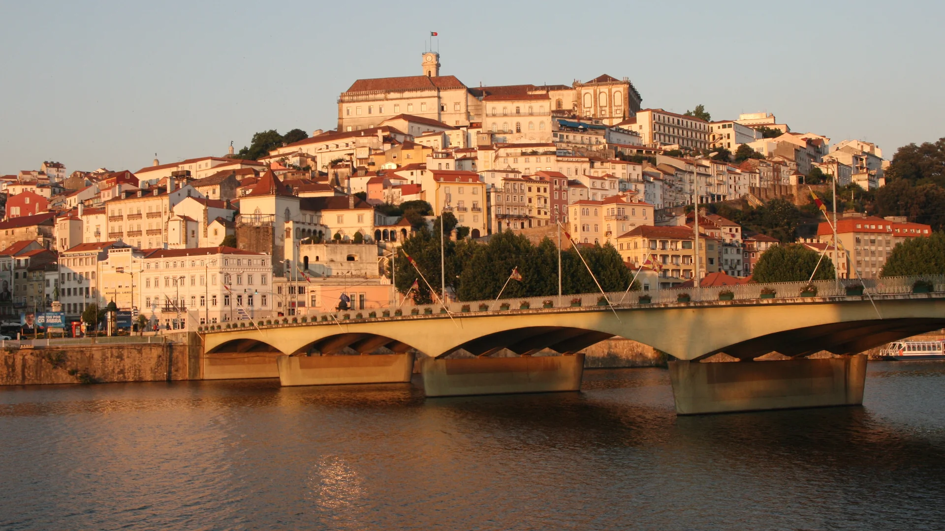Die Altstadt von Coimbra, die sich einen Hügel über einer Brücke über den Mondego hinaufzieht, ihre hellen Häuser und roten Dächer von der Abendsonne golden beleuchtet