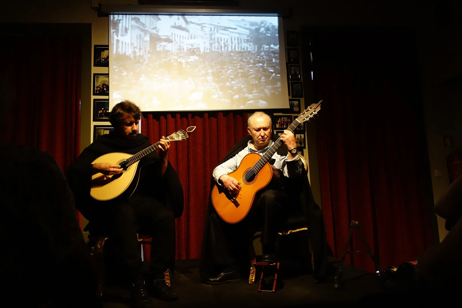 Zwei Musiker in schwarzen Umhängen sitzen auf einer kleinen Bühne, einer mit einer Coimbra-Gitarre und einer mit einer klassischen Gitarre, vor roten Vorhängen und einer Projektionsleinwand